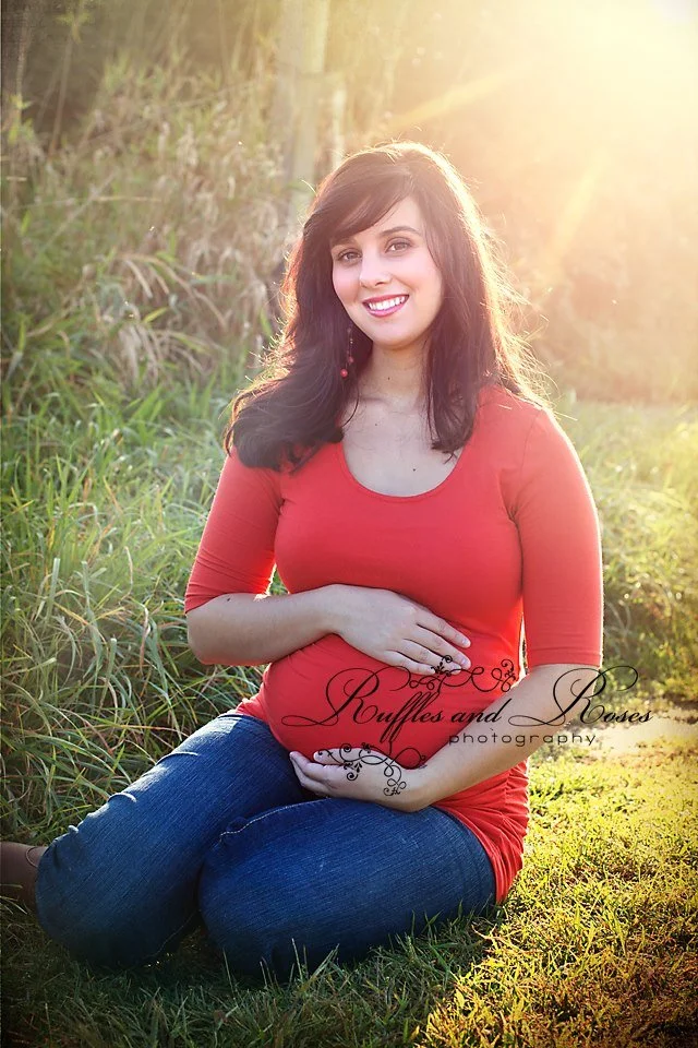 A pregnant woman with dark hair in a red top and blue jeans sitting on grass in a field during golden hour sunlight, smiling at the camera.
