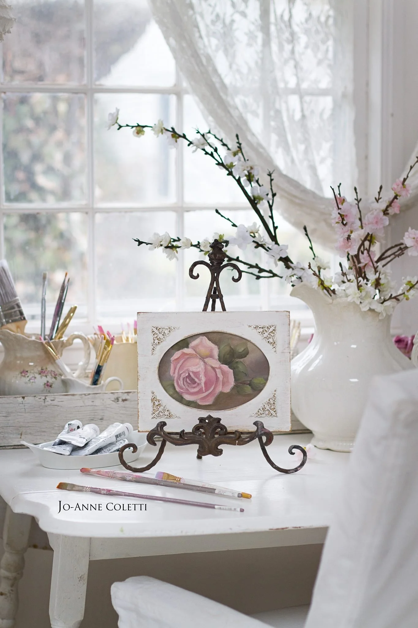 A white wooden table with art supplies including paint tubes, brushes, and a palette. A framed painting of pink roses on a stand. Behind, a window with lace curtains and cone-shaped trees outside.