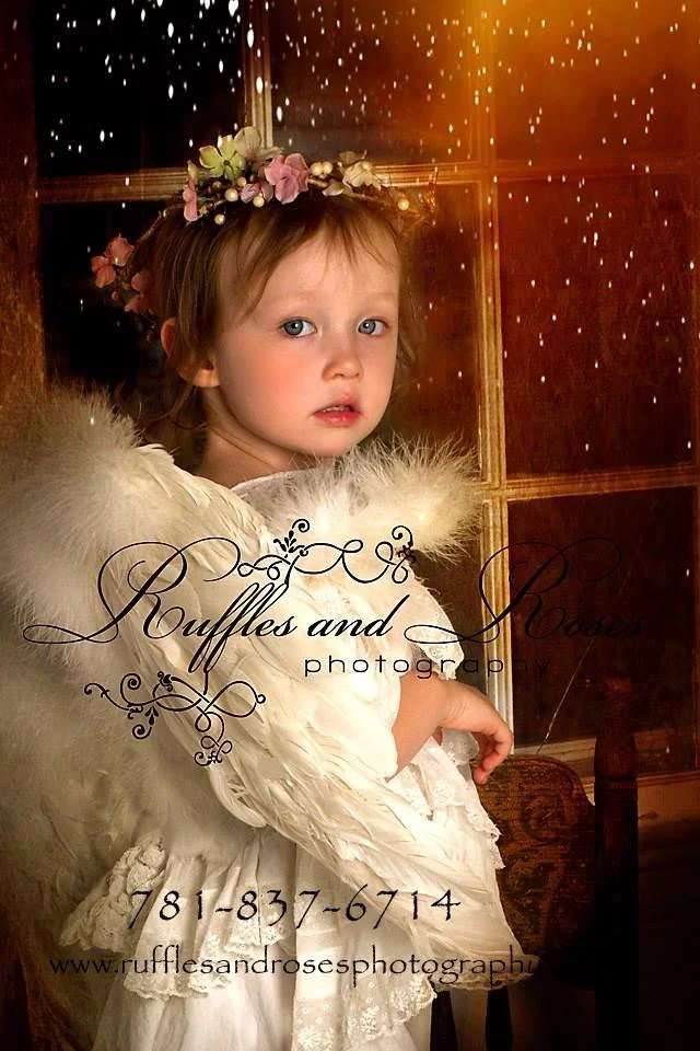 A young girl with a floral headband and blue eyes, dressed in a white dress with a fluffy shawl, standing indoors in front of a wooden paneled background with a window.
