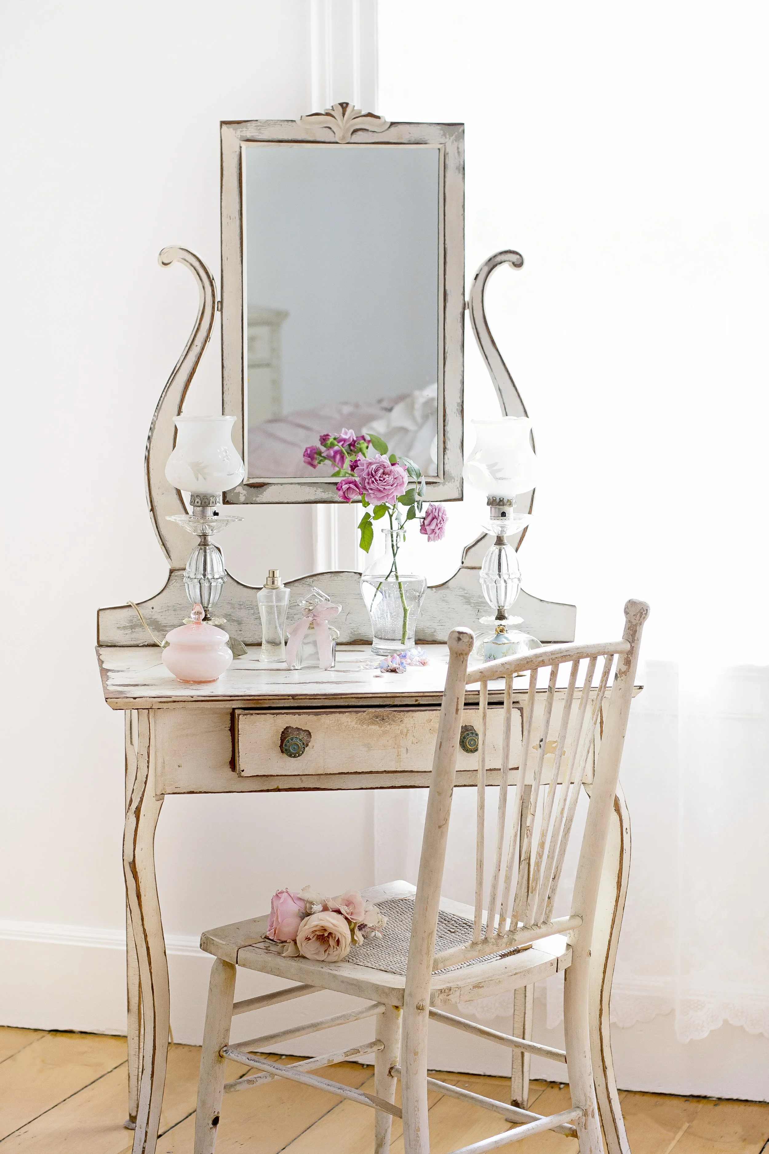 Vintage dressing table with a mirror, pink flowers in a glass vase, and various perfume bottles, paired with a distressed white wooden chair in a bright, cozy room.