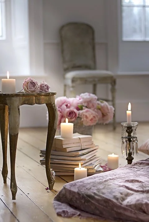 A cozy room decorated with pink roses, lit candles, and soft pillows, featuring a vintage chair and a small wooden table.