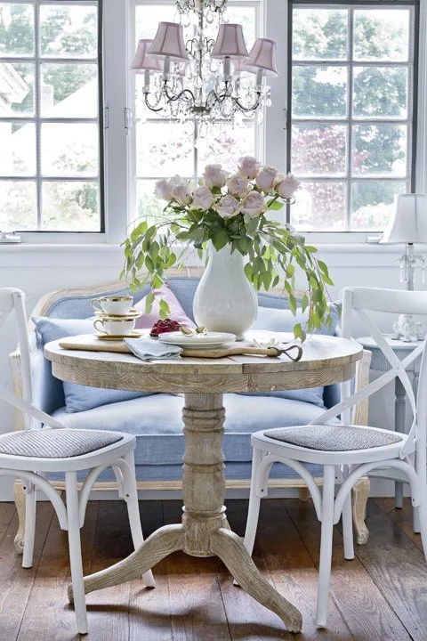 A cozy dining area with a round wooden table, white chairs, and a blue upholstered bench. The table has a white pitcher with pink roses and green leaves, teacups, and saucers. A chandelier with pink lampshades hangs above, and windows in the backgrou