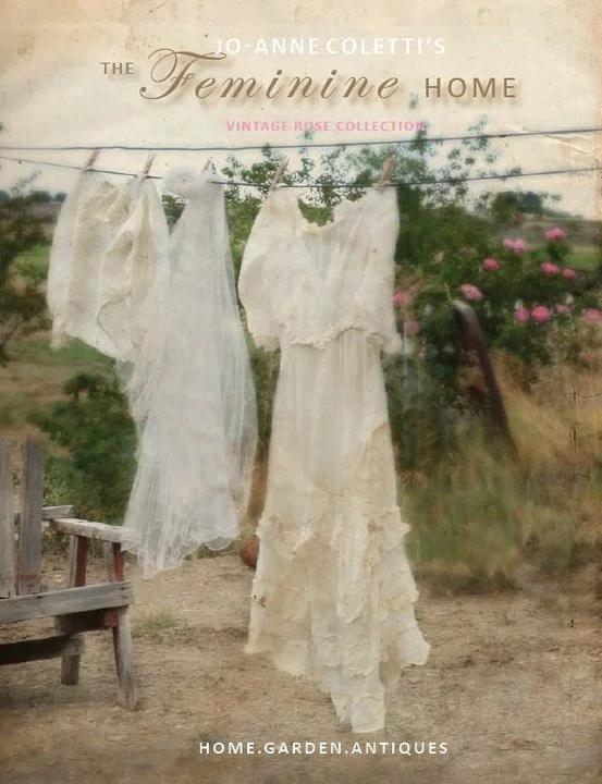 Vintage white dresses or fabric hanging on a clothesline outdoors. A rustic wooden bench is visible on the left, and greenery with pink flowers in the background.