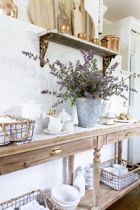 Wooden table with white dishes, a pitcher, and a large flower pot with purple flowers, against a white brick wall with a decorative shelf holding cutting boards and copper pots.