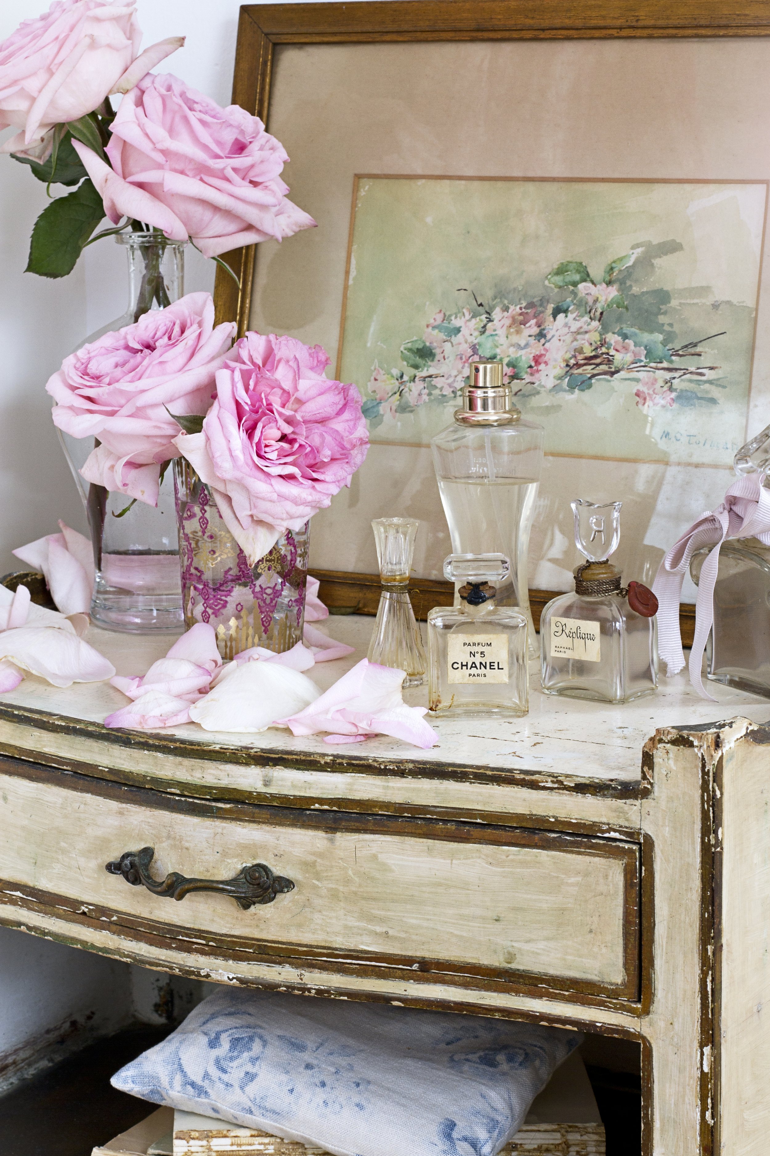 A vintage wooden dresser with a distressed white finish, displaying pink roses in a glass vase, perfume bottles, and a framed watercolor painting of flowers.