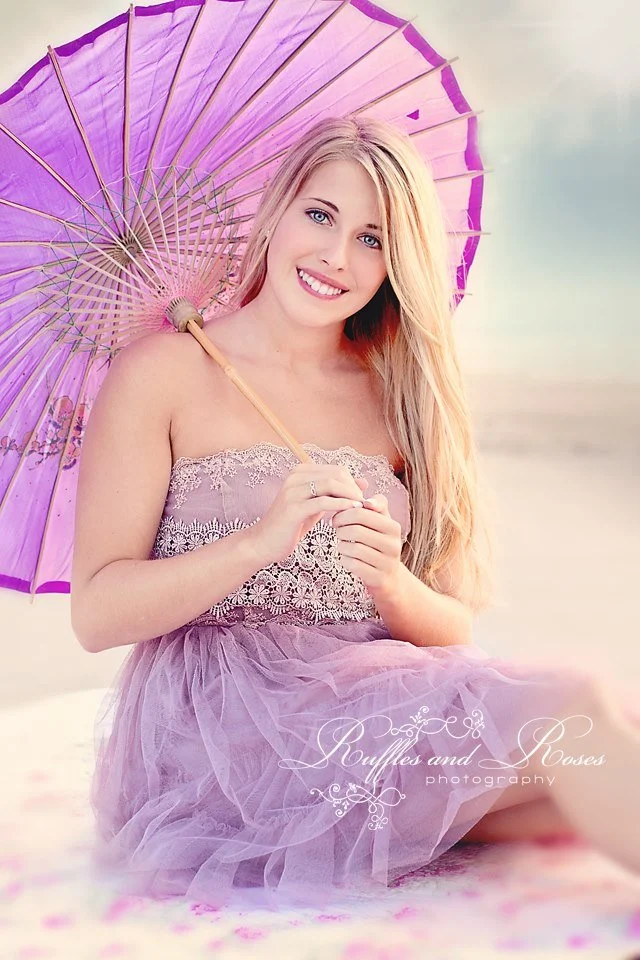 A smiling young woman with long blonde hair holding a purple parasol, sitting outdoors near a body of water, wearing a purple dress with lace details.