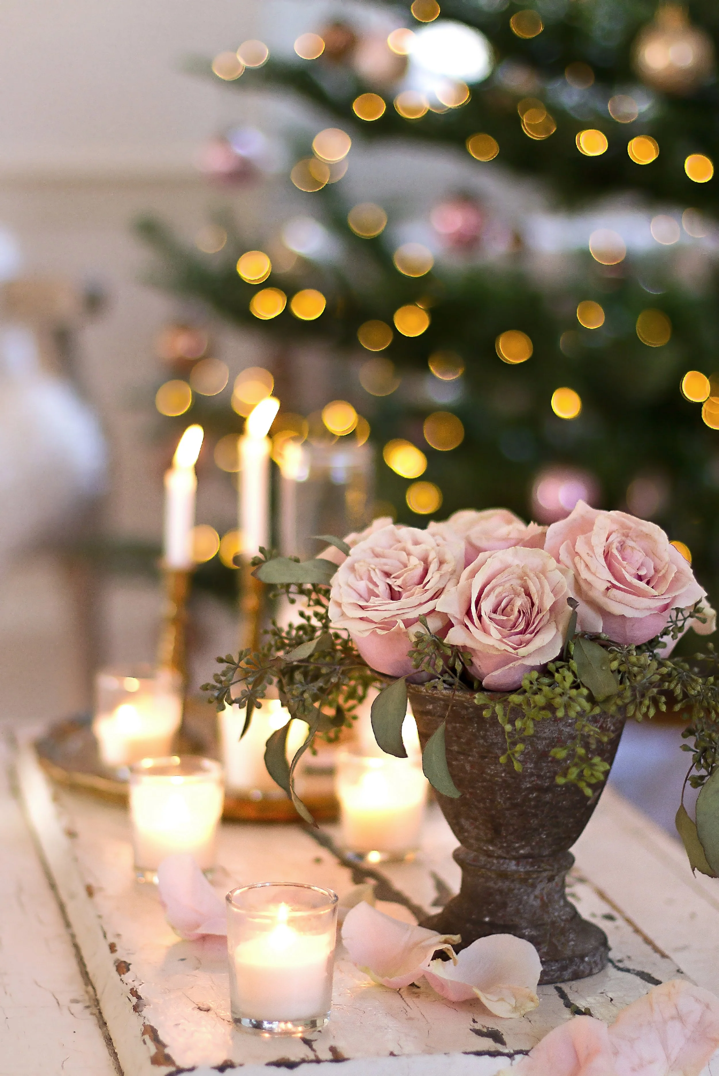 A rustic vase filled with soft pink roses and green foliage, surrounded by glowing candles and scattered rose petals, with a blurred decorated Christmas tree with lights in the background.