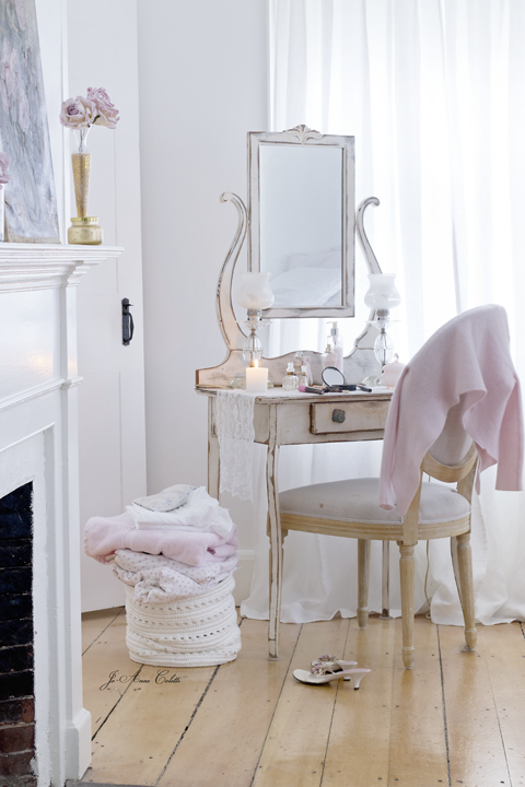 A vanity table with a mirror, candles, and glass bottles, with a pink garment draped over a chair, a pile of blankets on a woven basket, and a high-heeled shoe on the wooden floor.