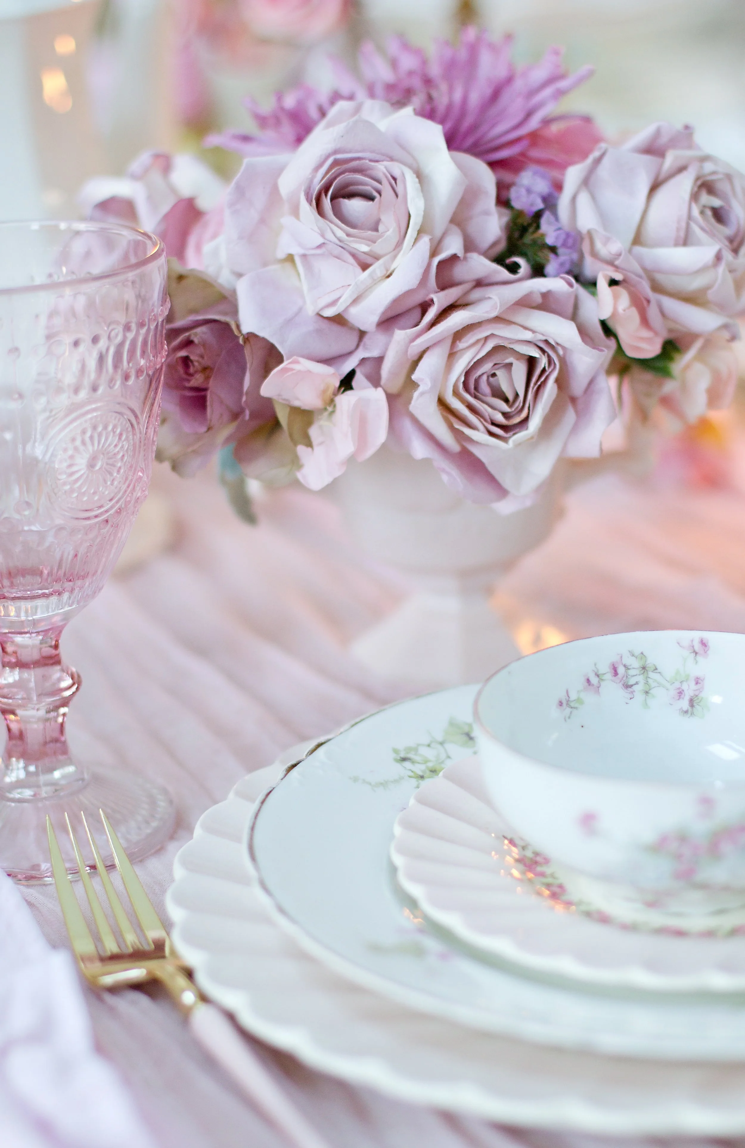 A close-up of a formal table setting with pink flowers in the background, delicate china with floral patterns, gold cutlery, and a pink glass drinking vessel.