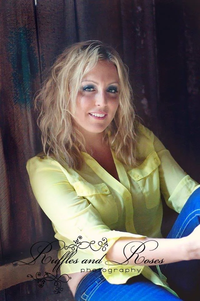 A woman with blonde, wavy hair wearing a yellow blouse and blue jeans, sitting against a wooden wall.