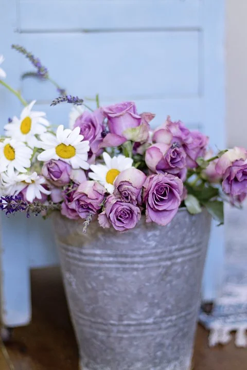 A bouquet of pink roses and white daisies with yellow centers in a gray pot with a textured surface, against a blue wooden background.