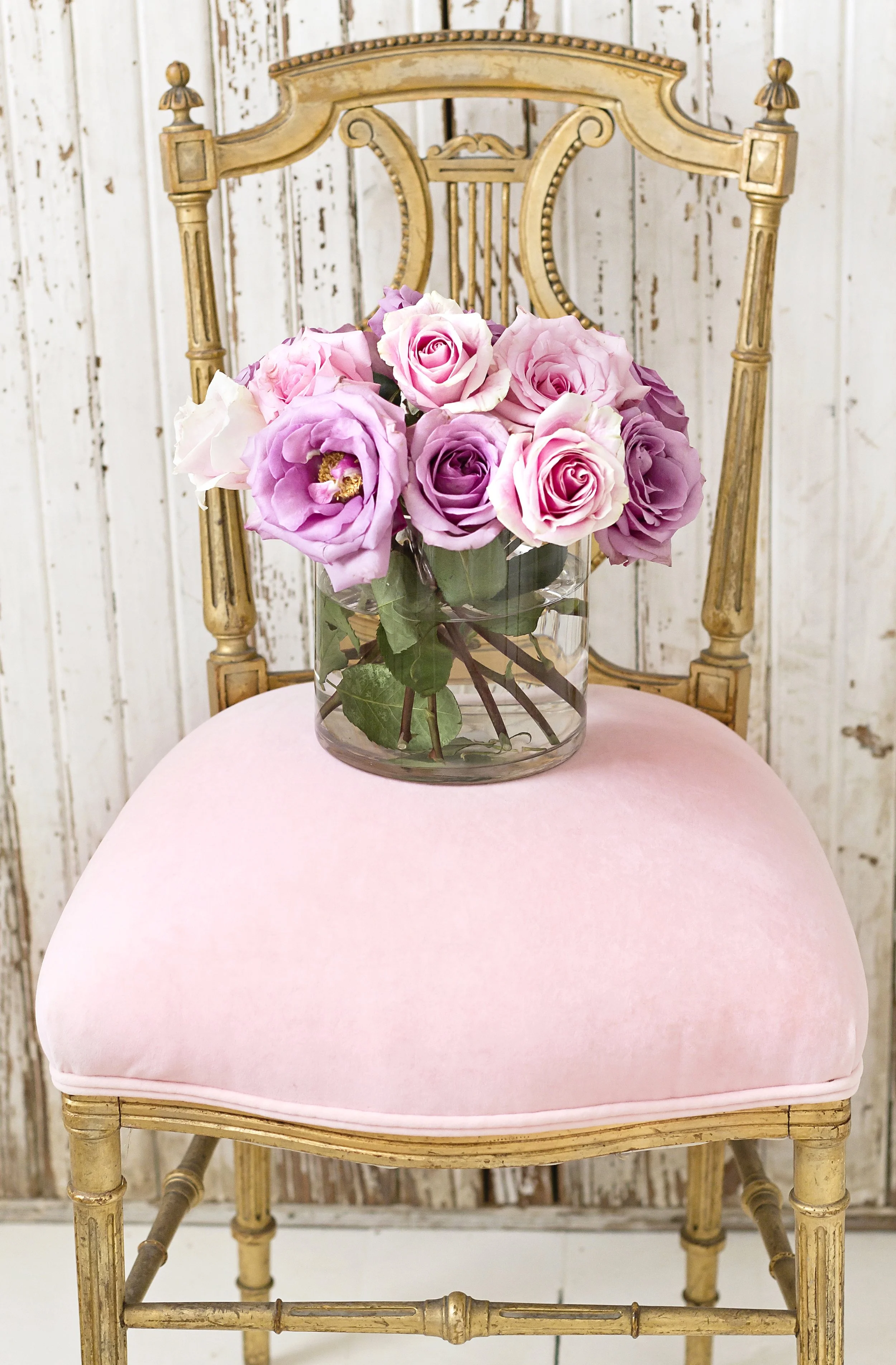 A glass vase of pink and purple roses on a pink upholstered chair with a gold ornate frame, in front of a rustic white wooden background.