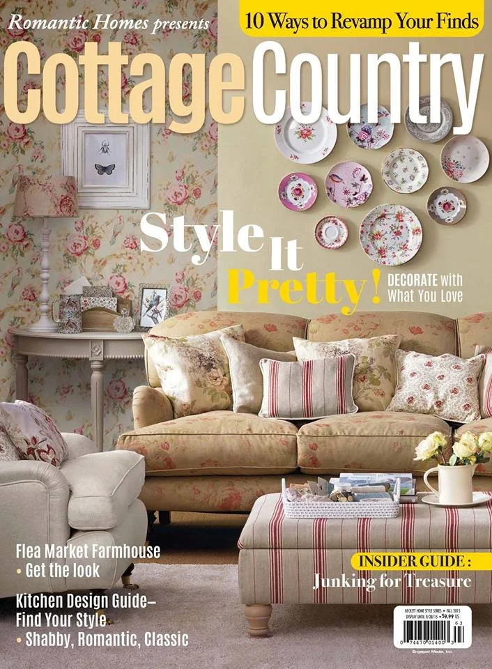 Living room interior with floral wallpaper, a floral sofa, striped and floral throw pillows, a small armchair, a table with a striped tablecloth, and a wall decorated with floral plates.