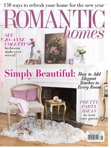 Cover of Romantic Homes magazine featuring a cozy living room with a pink armchair, a small gold table with pink flowers, a white fluffy rug, and a white fireplace with vases of flowers and a painting above it.
