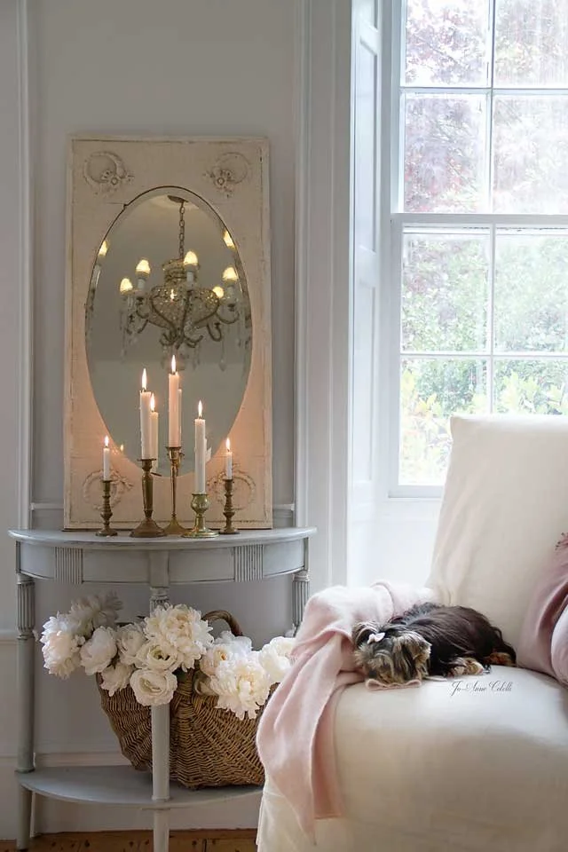 A cozy corner with a white sofa, a pink blanket, and a small striped tabby kitten sleeping. Behind the sofa is a large window with white framing, showing trees outside. To the left is a vintage white console table with a basket of white flowers and a
