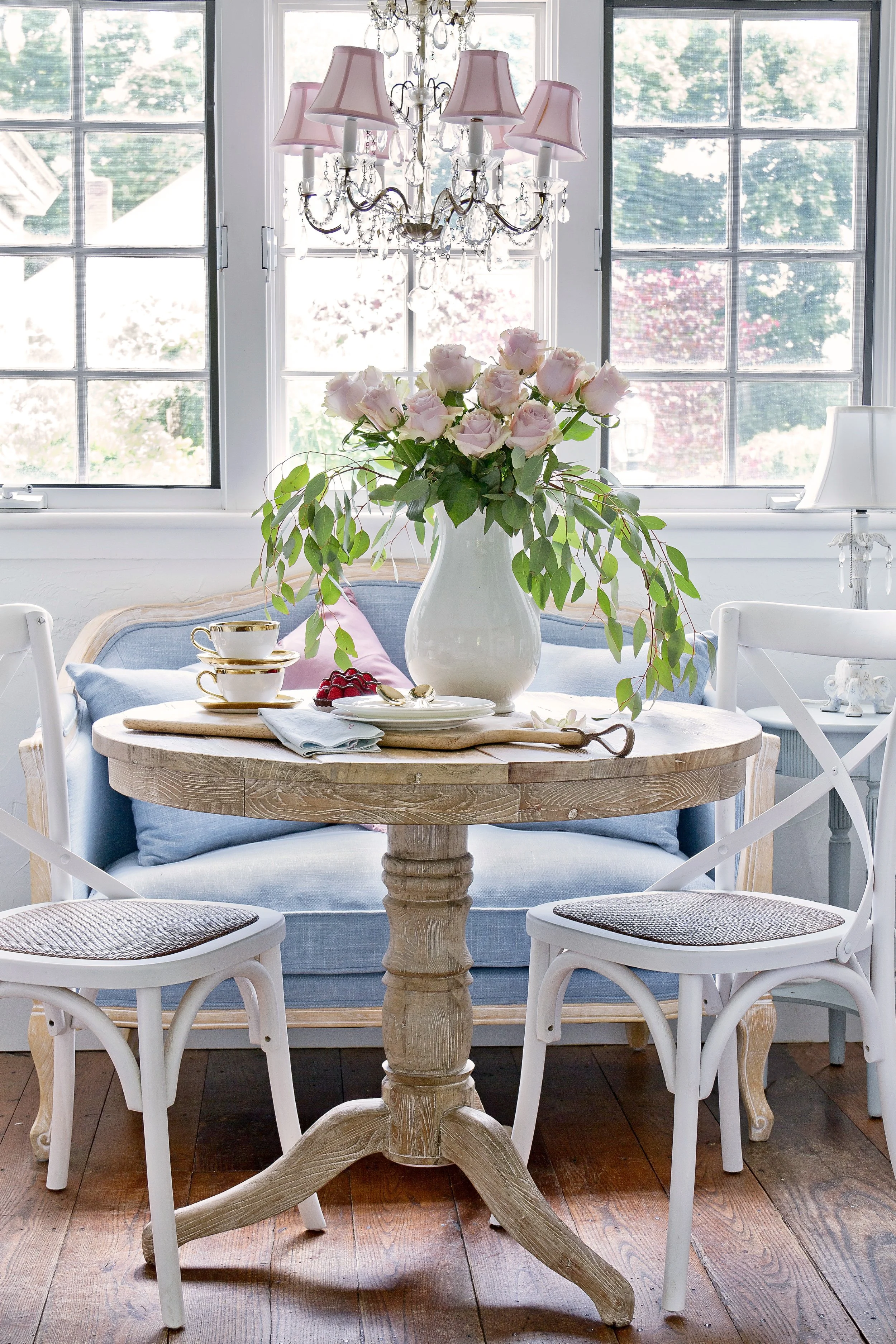 Round wooden dining table with white chairs, a large white vase filled with pink roses, a stack of white plates, a teacup set, and a small dish with berries, in a bright room with large windows and a chandelier.