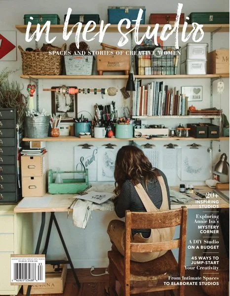 A woman with long brown hair in a striped shirt working at a cluttered desk in a cozy, creatively decorated studio with shelves filled with books, baskets, and art supplies.