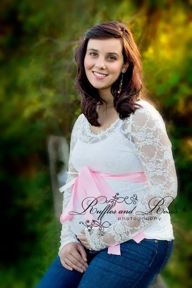 Young woman with dark brown hair and a white lace top, sitting outdoors in a natural setting with greenery, smiling at the camera.