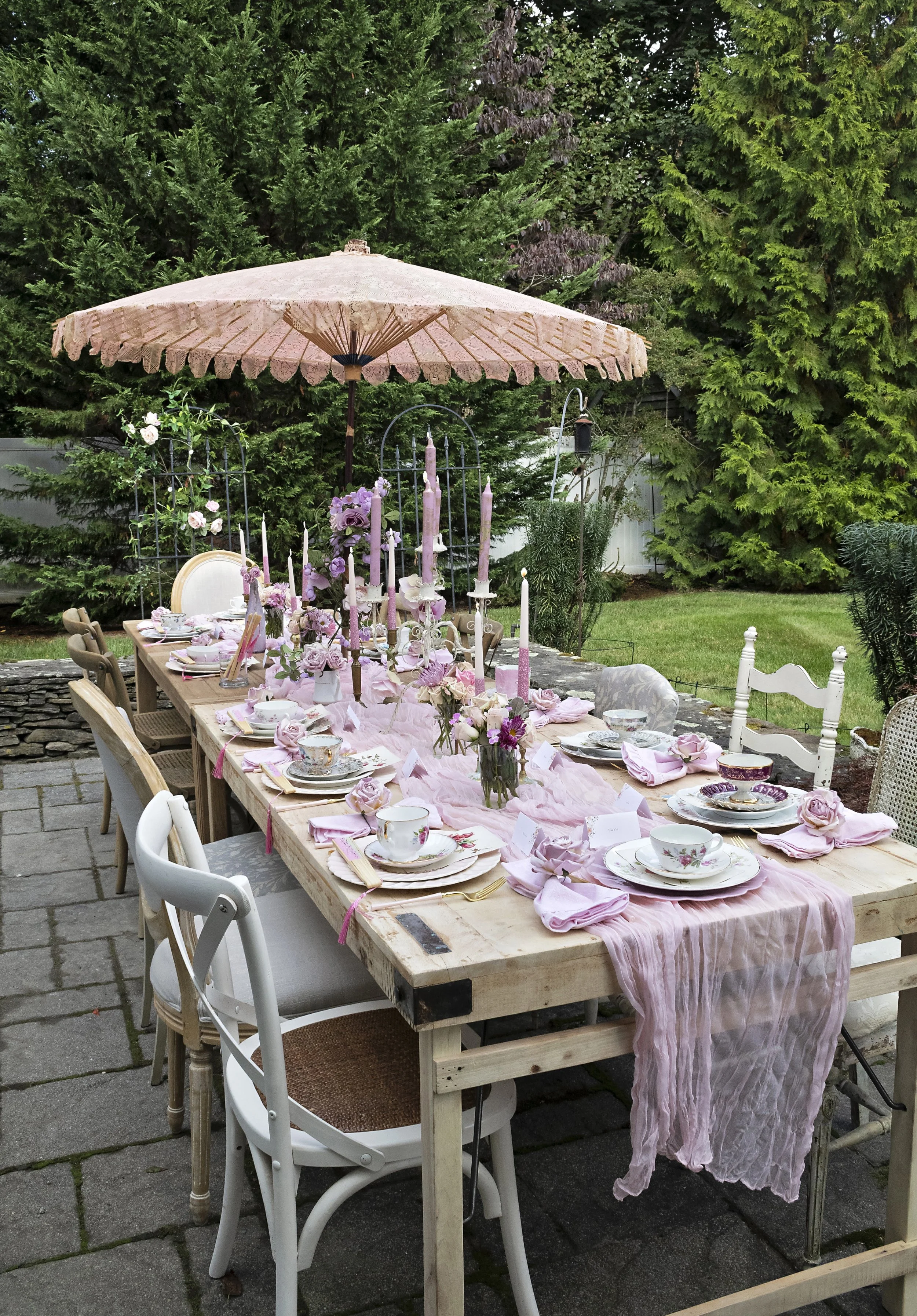 An outdoor dining table decorated with pink linens, floral arrangements, and candles, set under a large pink parasol, surrounded by chairs, on a stone patio with lush green trees in the background.
