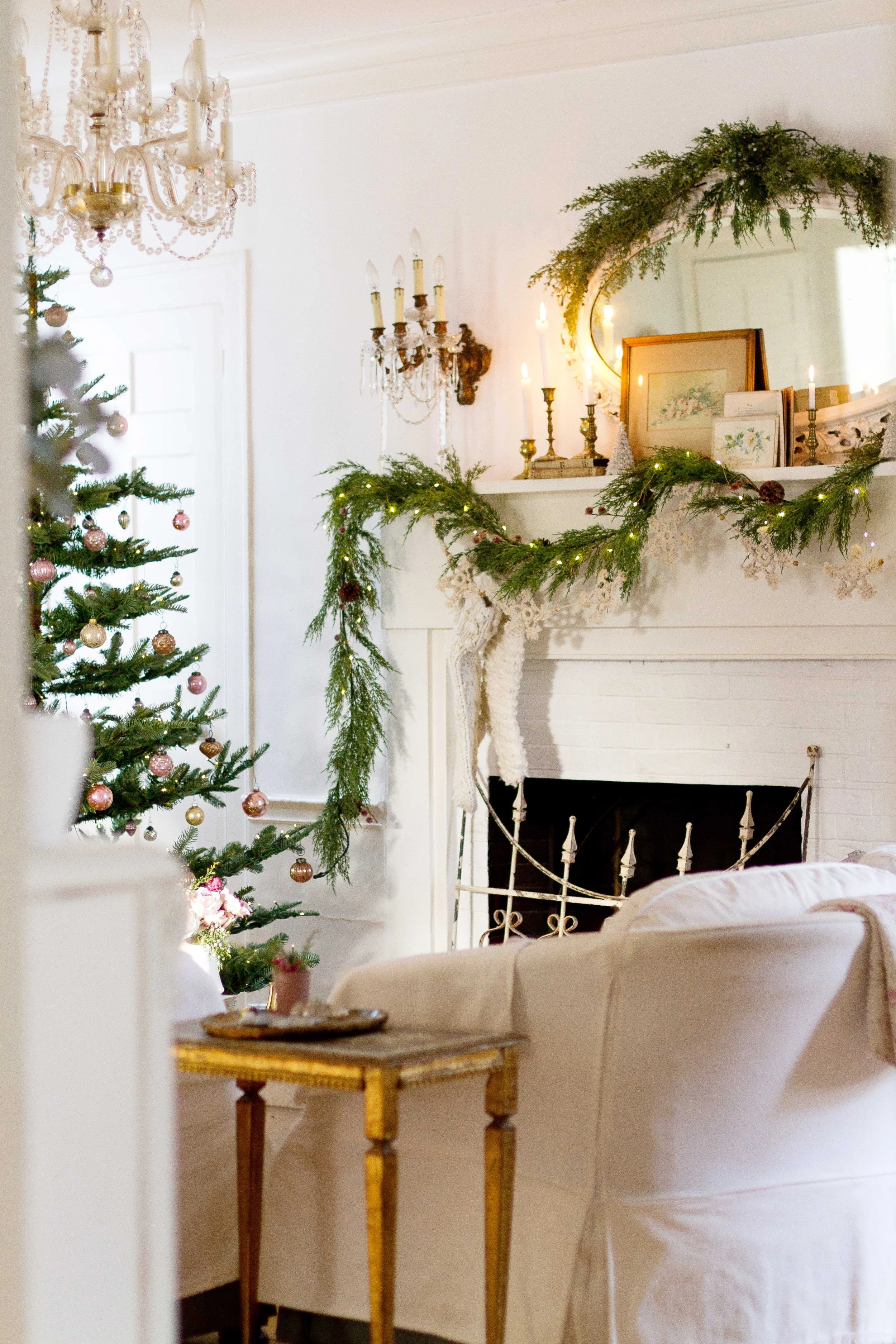 A cozy living room decorated for Christmas with a Christmas tree, garlands, candles, and framed pictures on a shelf, all in a white and soft color scheme.