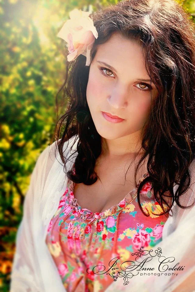 Portrait of a young woman with dark, wavy hair wearing a floral dress and a light cardigan, outdoors with greenery in the background.