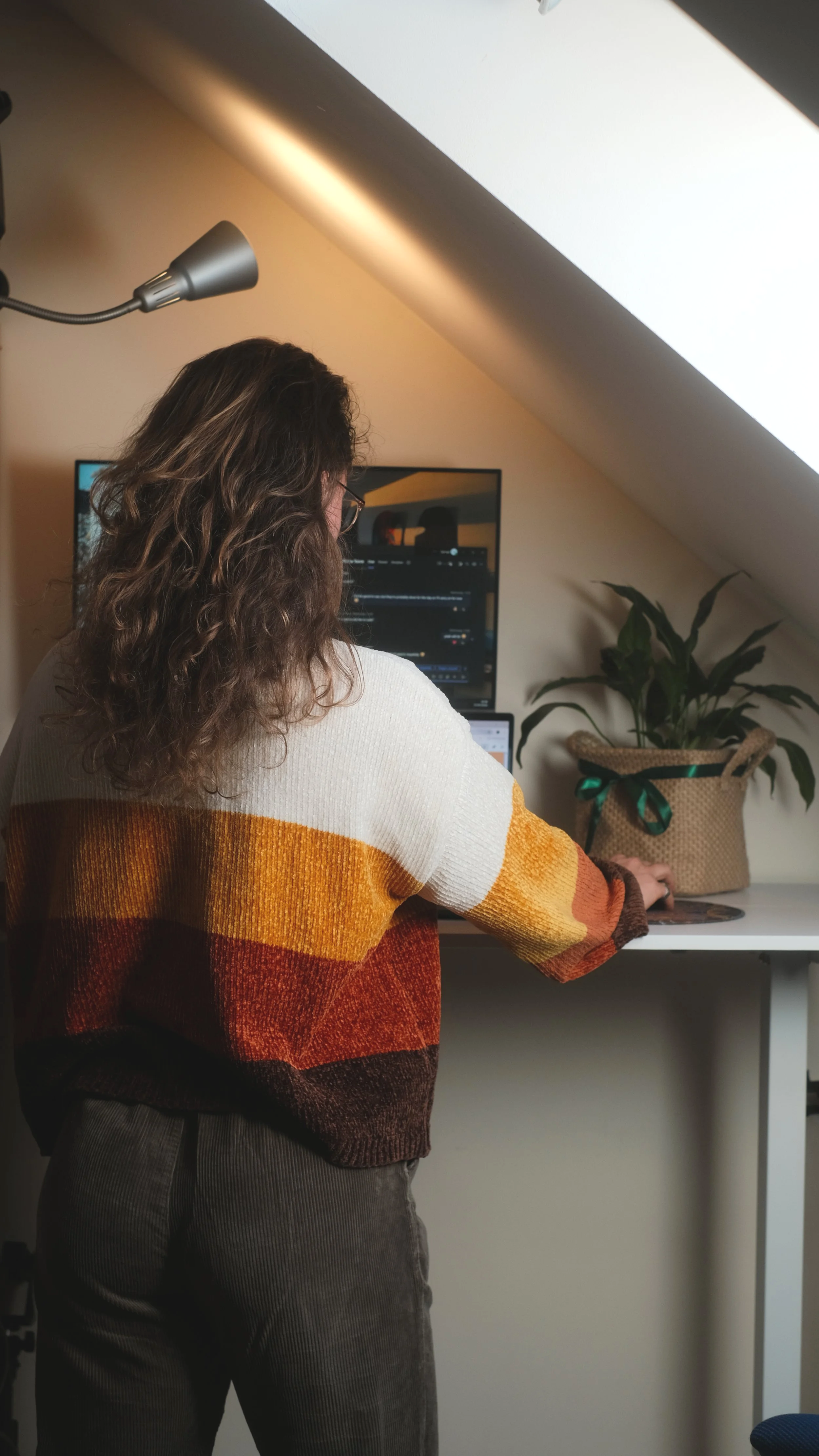 A woman with curly hair wearing a multicolored striped sweater working at a desk with a computer monitor and a potted plant in a woven container.