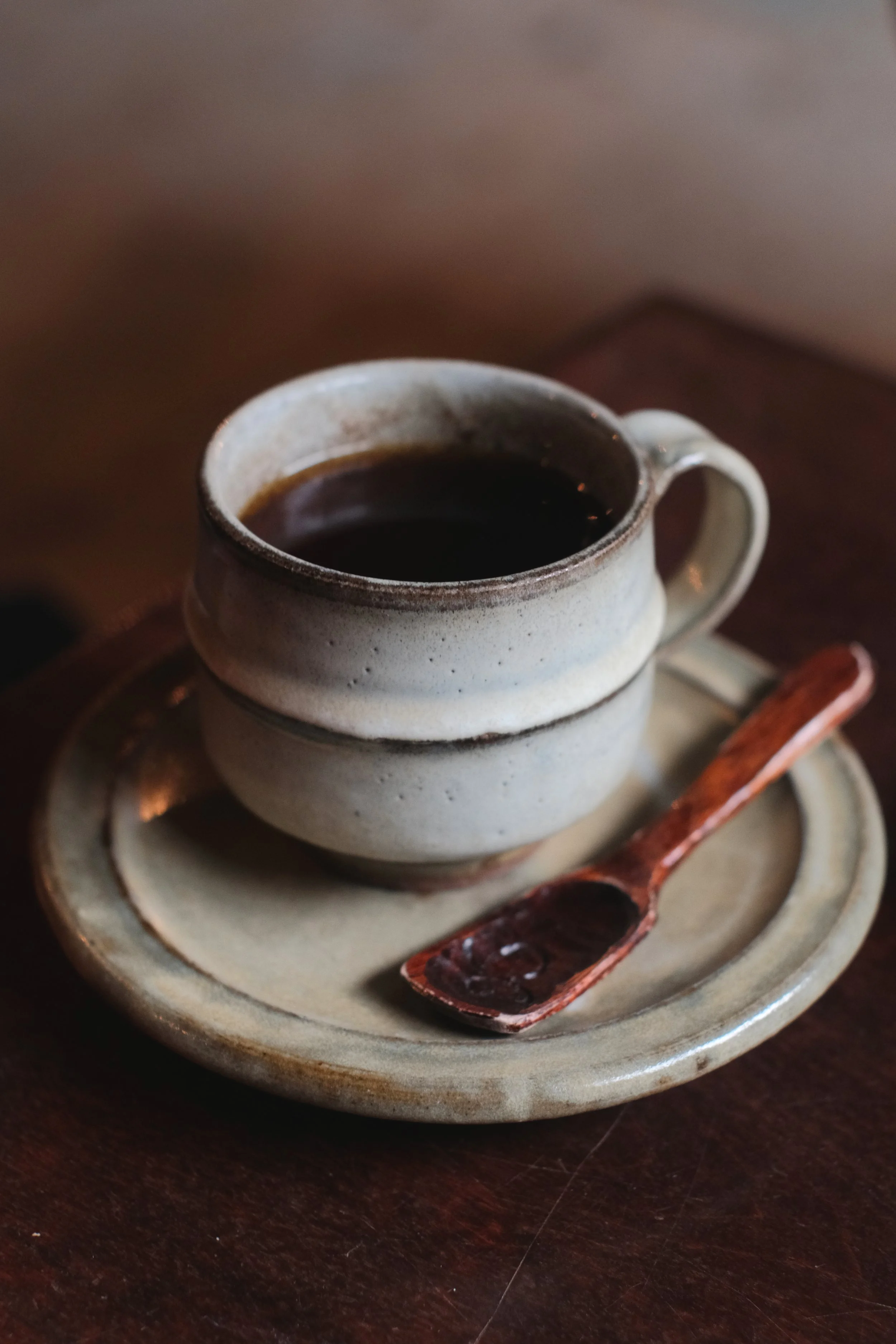 A ceramic cup filled with black coffee placed on a matching saucer, with a small copper spoon resting on the saucer.