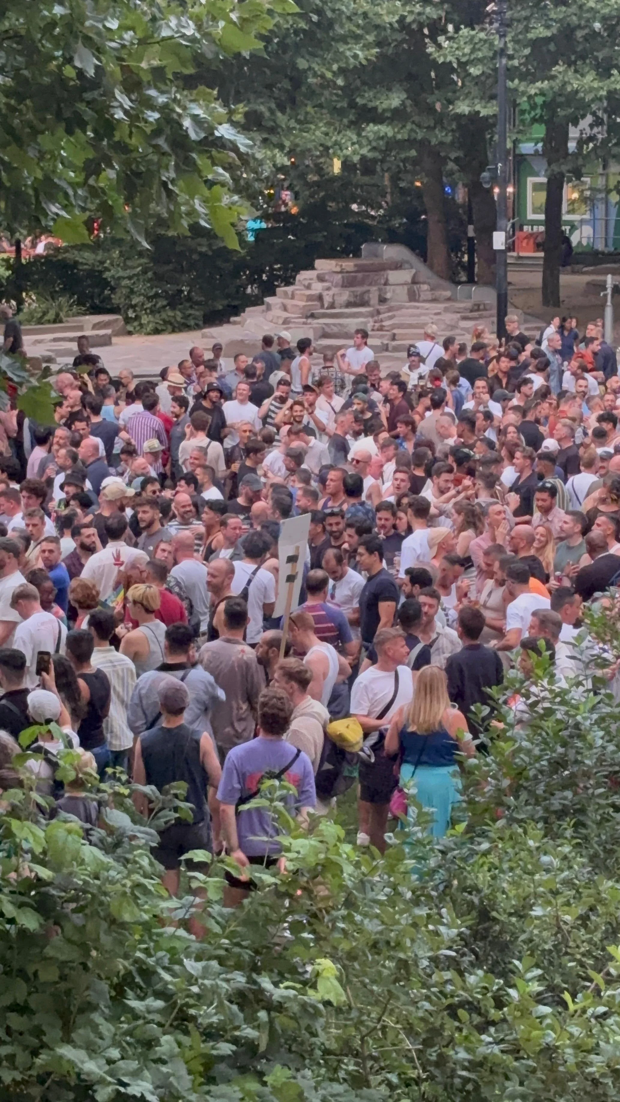 Large crowd of people gathered in an outdoor park surrounded by trees.