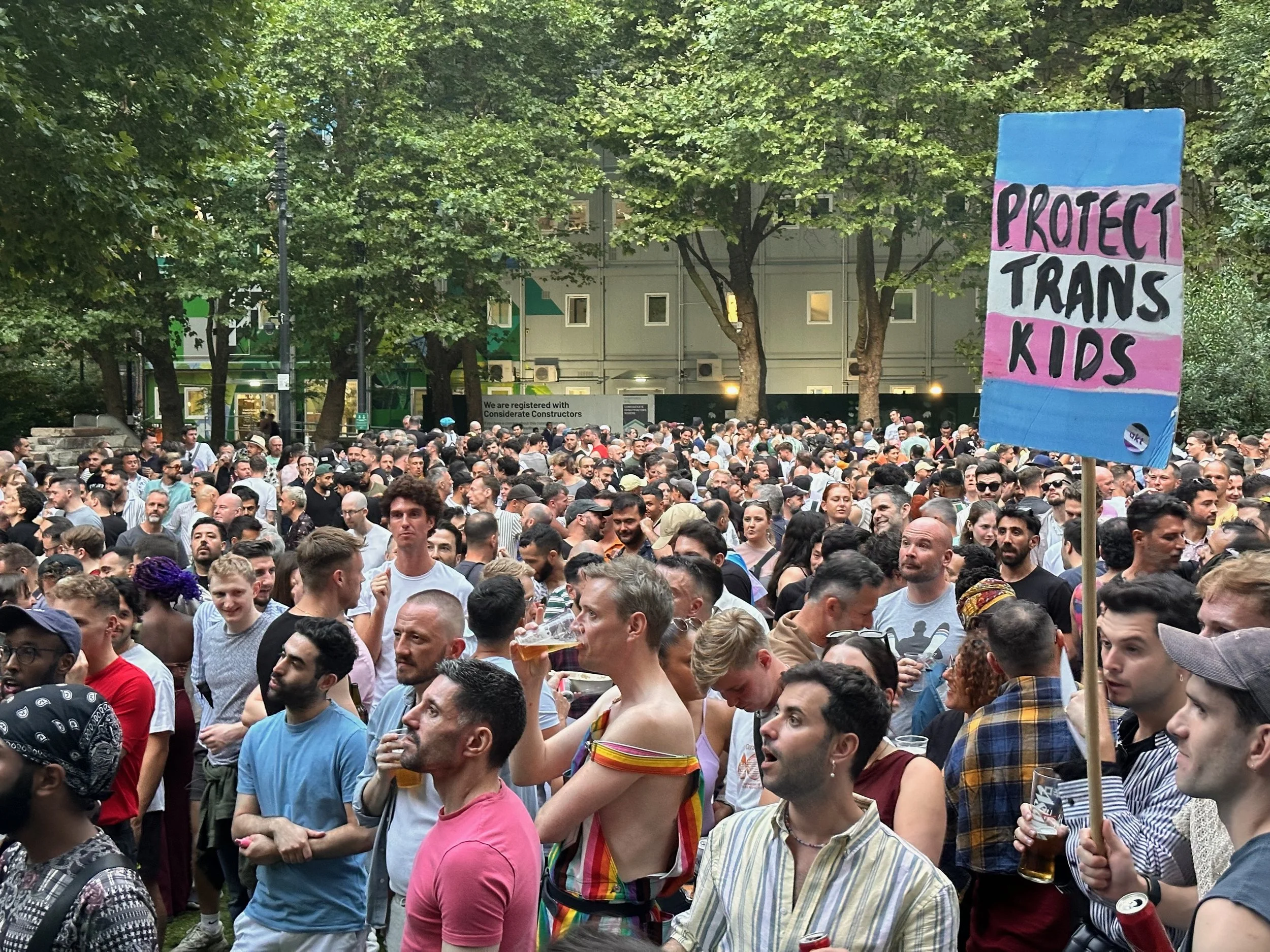A large crowd of diverse people gathered outdoors in a park for a demonstration or rally. Some are holding drinks, and one person is holding a colorful sign that reads 'Protect Trans Kids.'