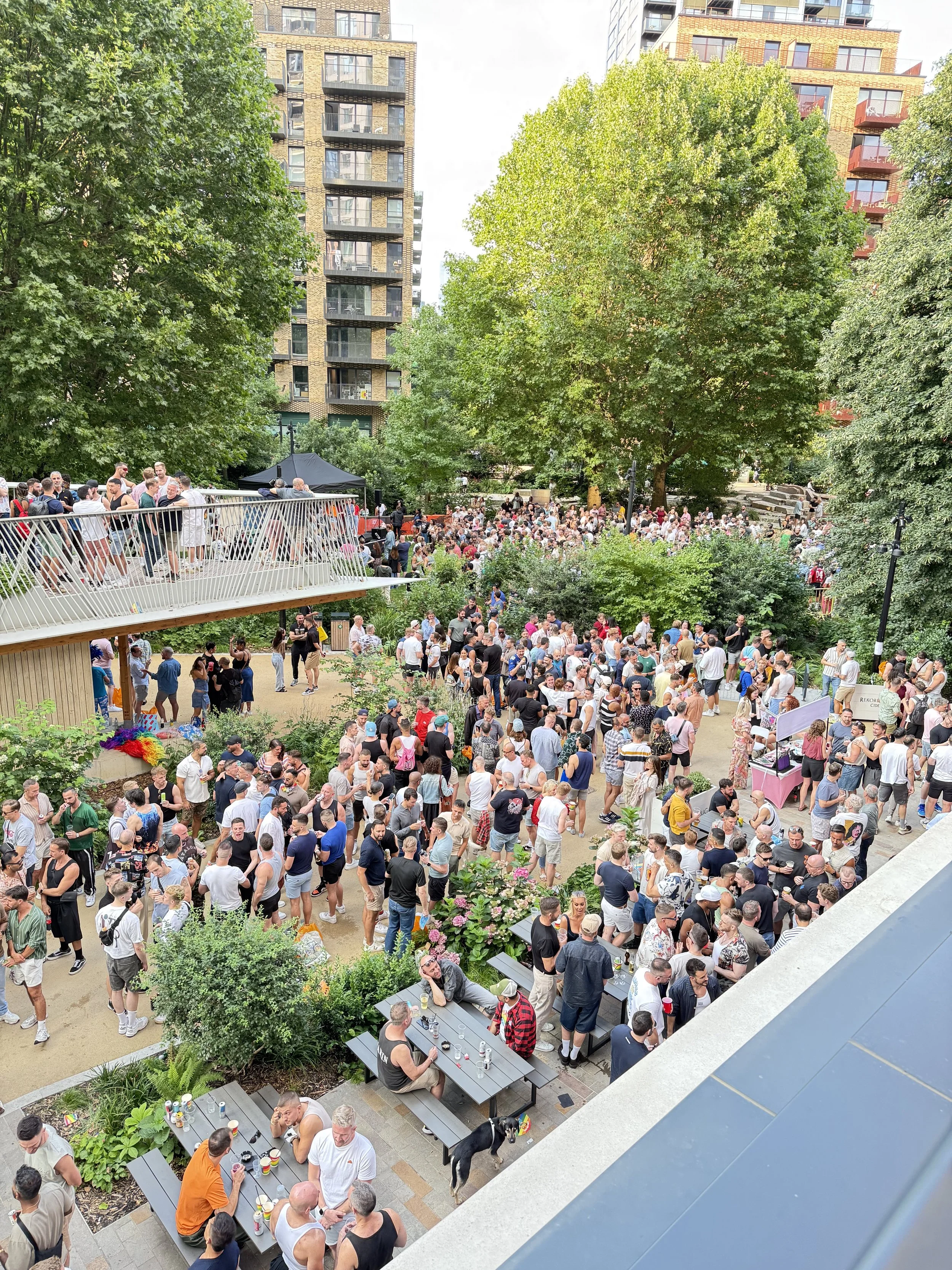 A crowded outdoor event in an urban park with many people socializing, with trees and high-rise apartment buildings in the background.