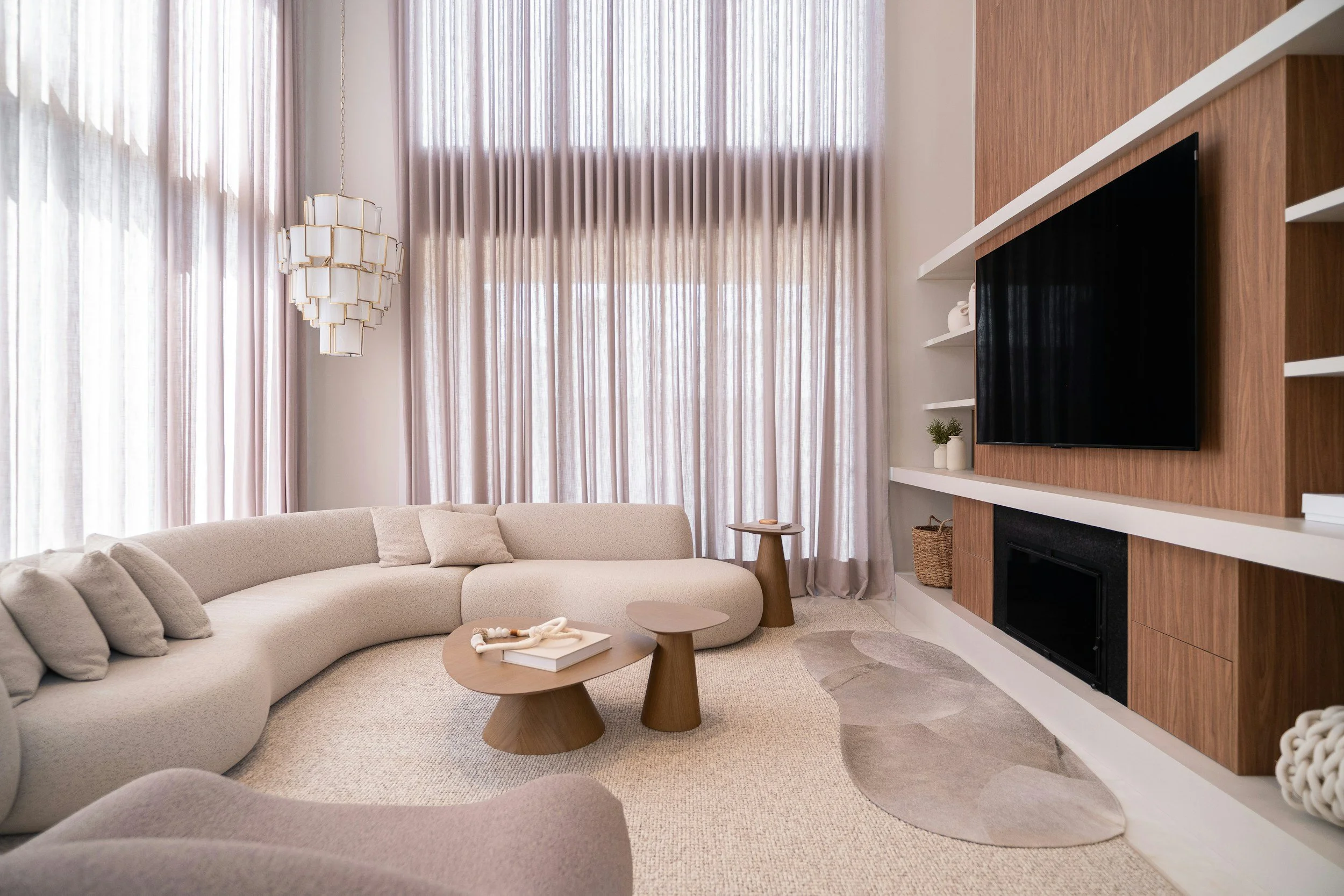 Living room with beige sectional sofa, two wooden coffee tables, tall window with sheer curtains, modern chandelier, wall with wood paneling, mounted flat-screen TV, white shelves with decorative items, fireplace, rug.