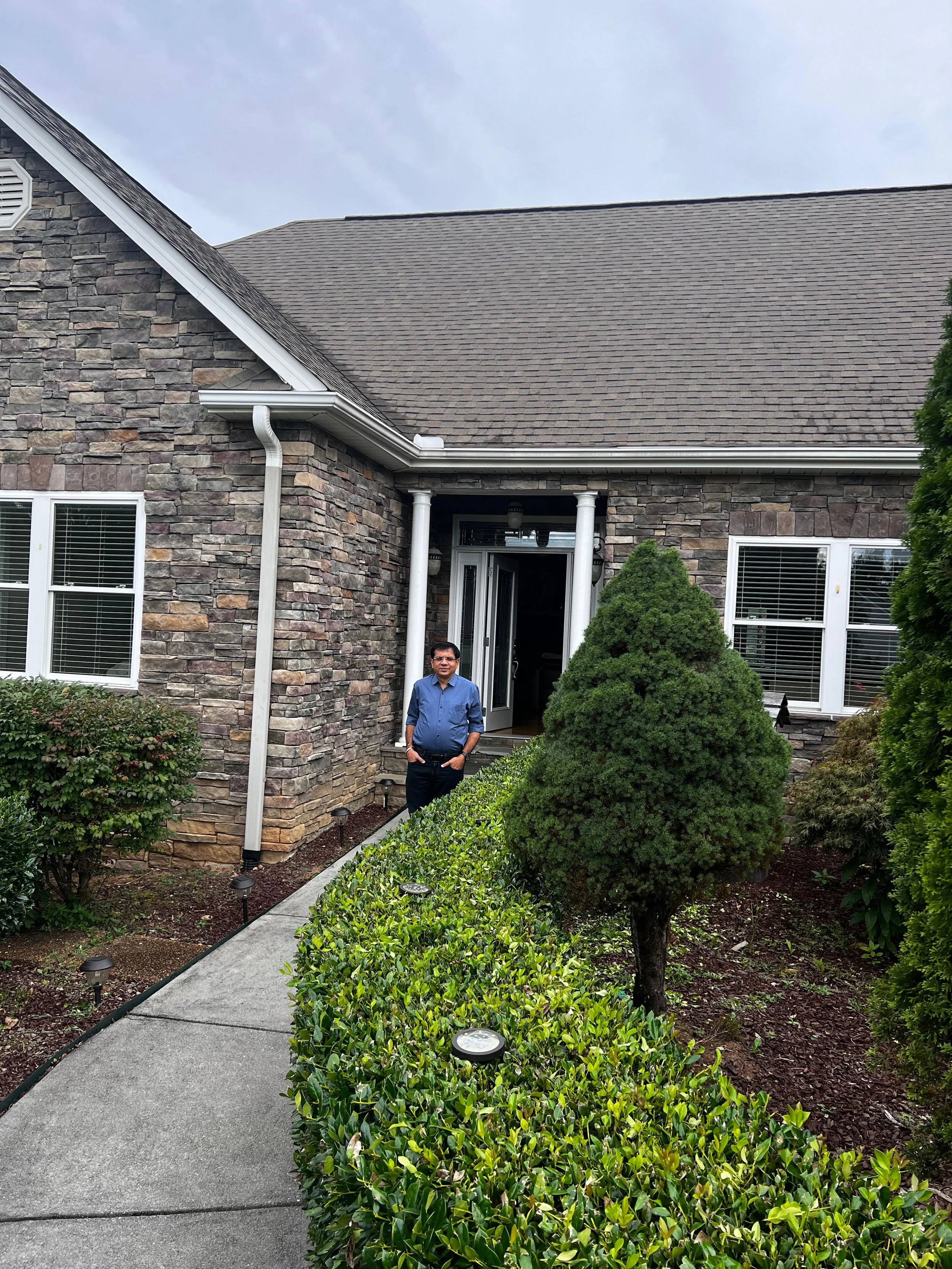 A man standing on a concrete sidewalk in front of a house with stone exterior walls and white window frames. The house has a peaked roof and a covered porch. There is a well-maintained garden with trimmed bushes, small trees, and landscape lights lining the path. The sky is overcast.
