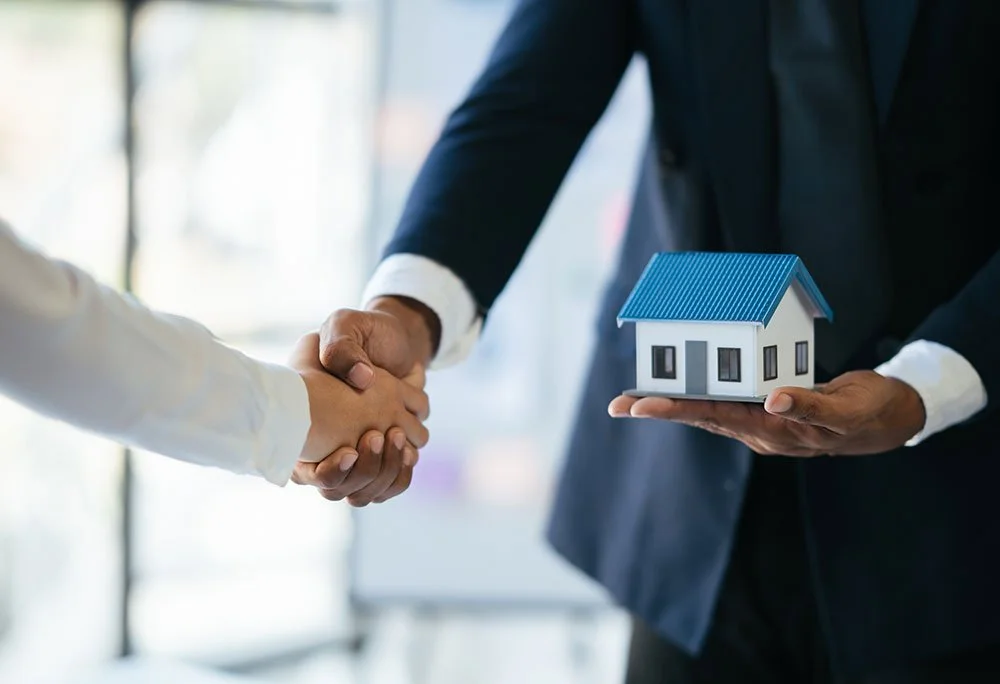 Person in a suit handing over a miniature house to another person during a handshake, in an indoor setting.