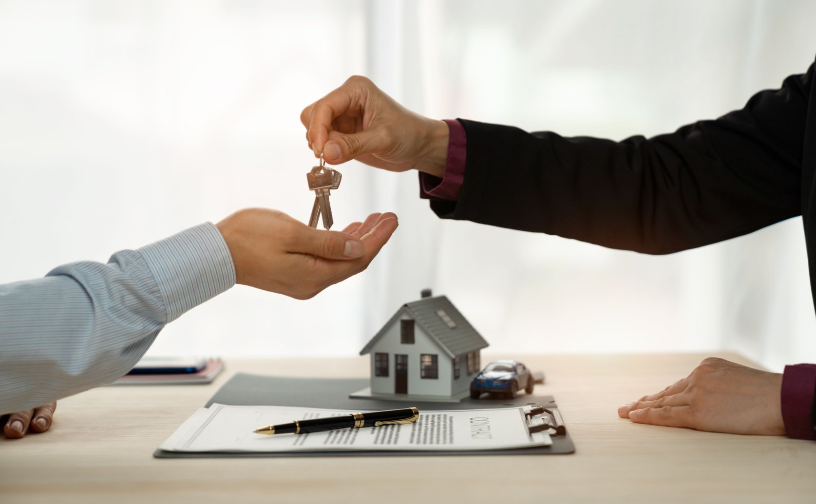 A person hands over house keys to another, with a model house and car on a desk. Nearby, a contract and pen lie on a clipboard, symbolizing a real estate deal
