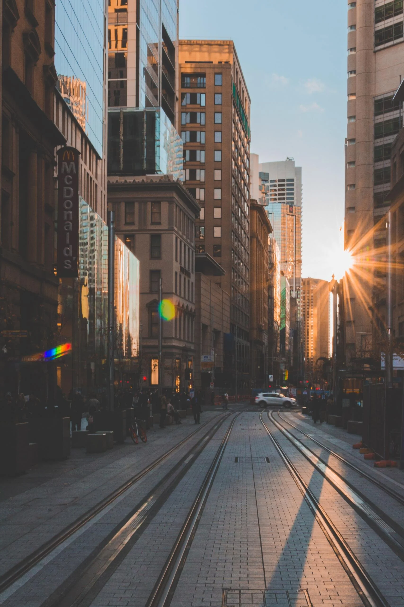 A city street featuring train tracks with buildings visible in the background under a clear blue sky.