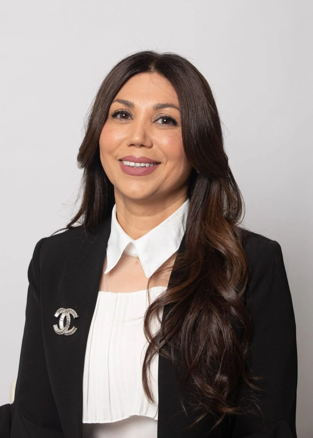 A professional woman with long brown hair, wearing a black blazer with a Chanel brooch, a white blouse, and smiling against a plain gray background.