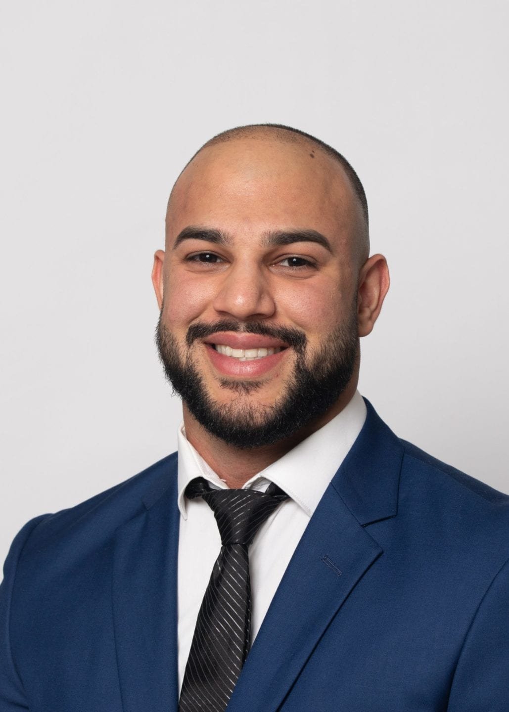 A man with a shaved head and a beard smiling, wearing a navy blue suit, white shirt, and black striped tie against a plain light gray background.