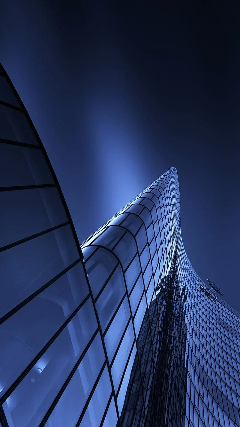 Looking up at a modern skyscraper with a curved glass facade at night, illuminated with blue lighting.
