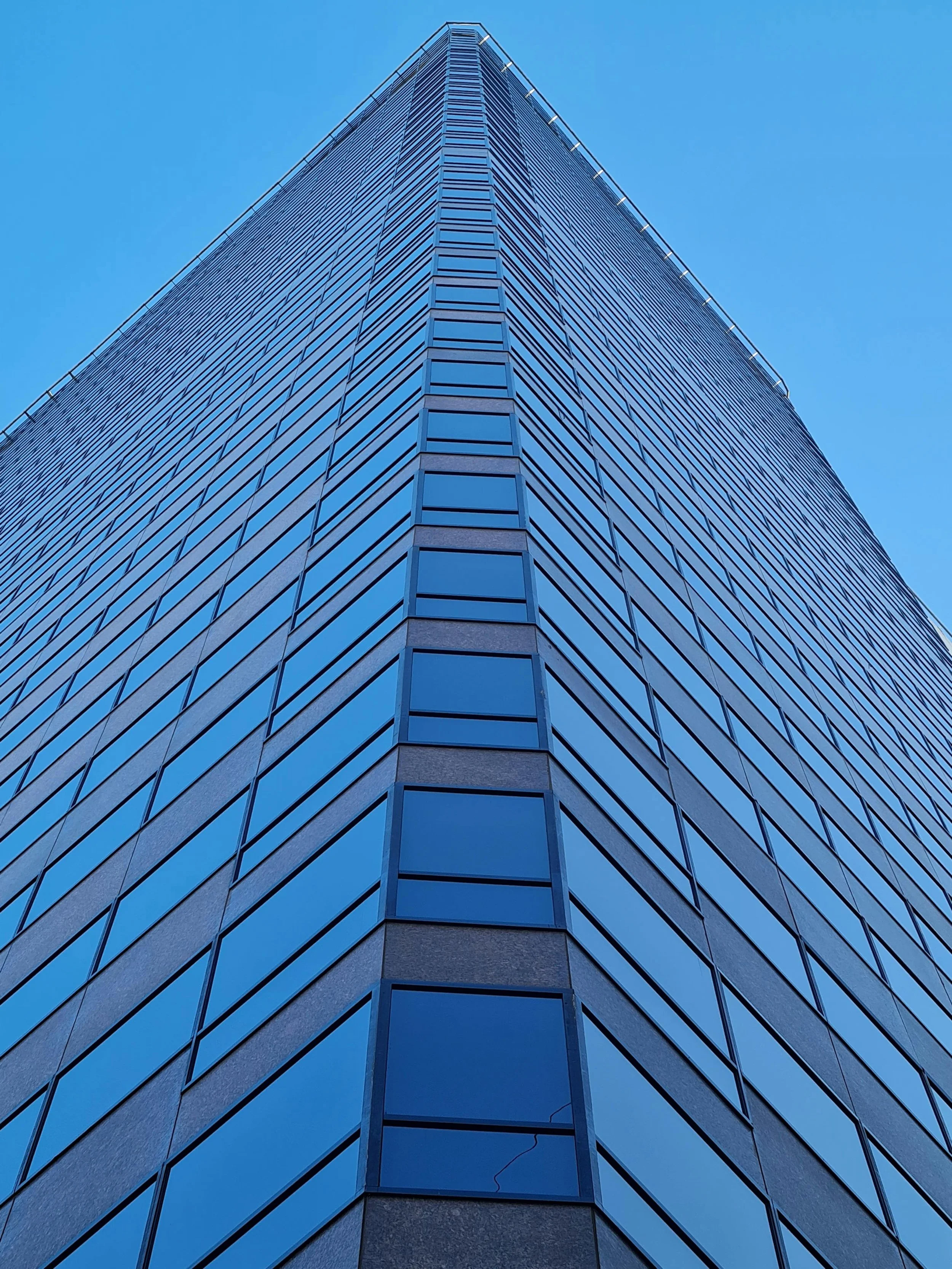 Upward view of a modern skyscraper's corner, with reflective blue glass windows and angular lines, set against a clear blue sky, conveying a sense of height