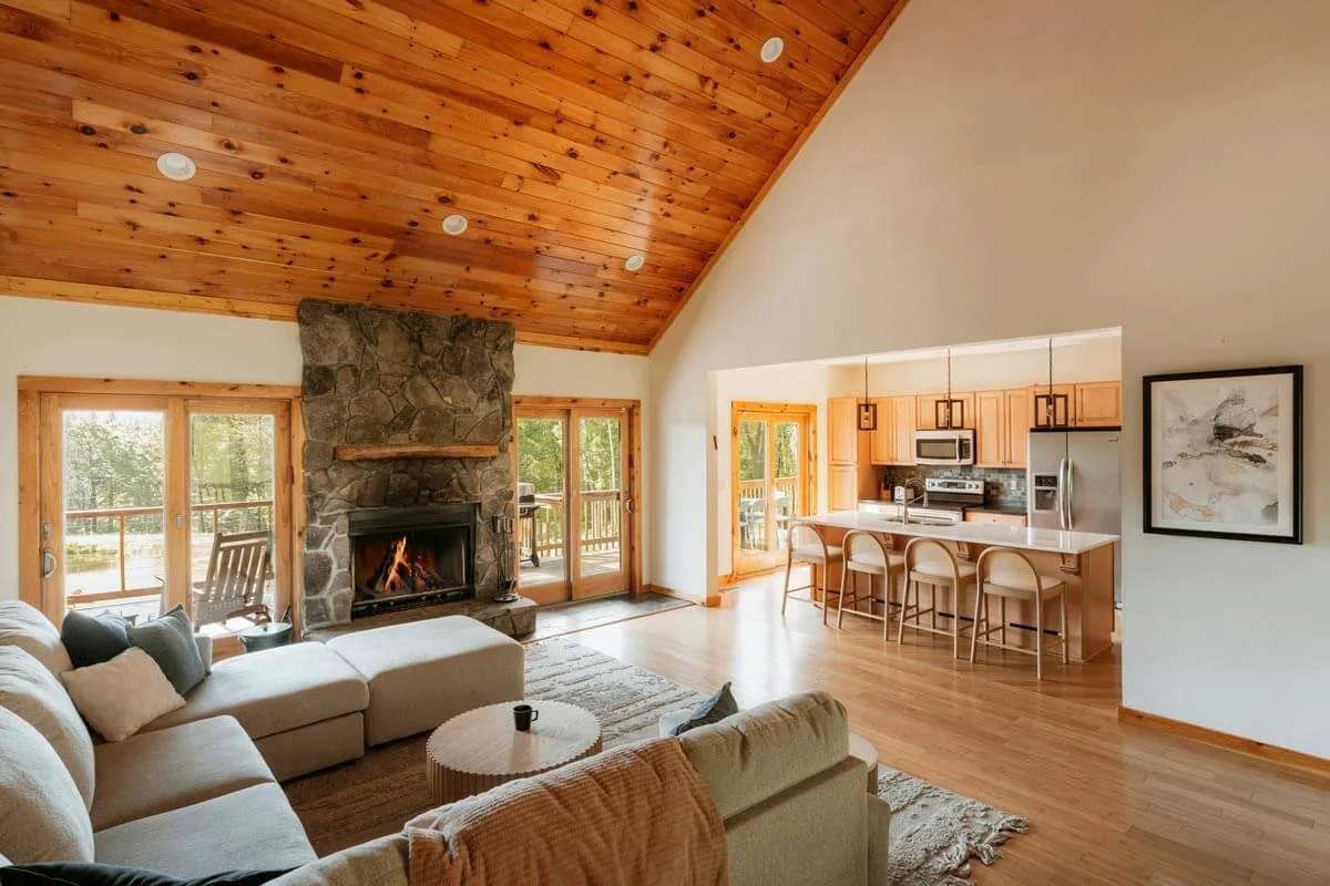 Cottage living room with a stone fireplace, beige sectional sofa, and wooden ceiling, open to a kitchen with wooden cabinets and countertops, and glass doors leading outside.