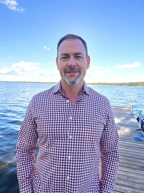 A man in a checkered shirt standing on a dock beside a lake with a blue sky overhead.