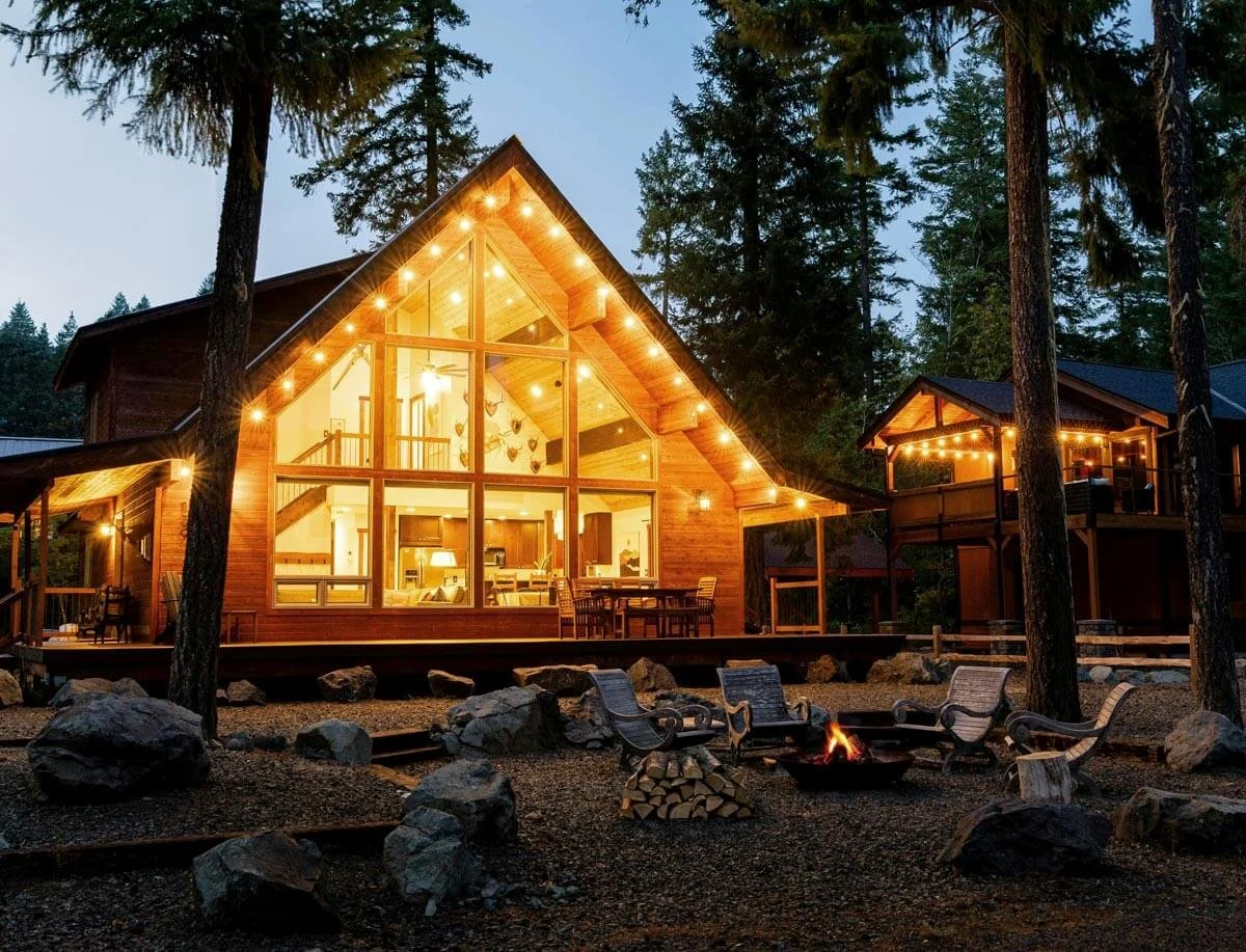 A large lake cottage with a triangular roof and extensive glass windows, illuminated by string lights, surrounded by tall trees and outdoor seating around a fire pit.