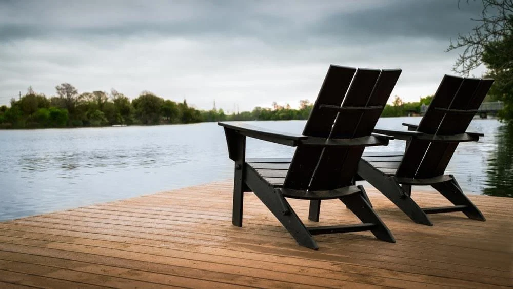Two black Adirondack chairs facing a lake on a wooden dock, with trees and cloudy sky in the background.