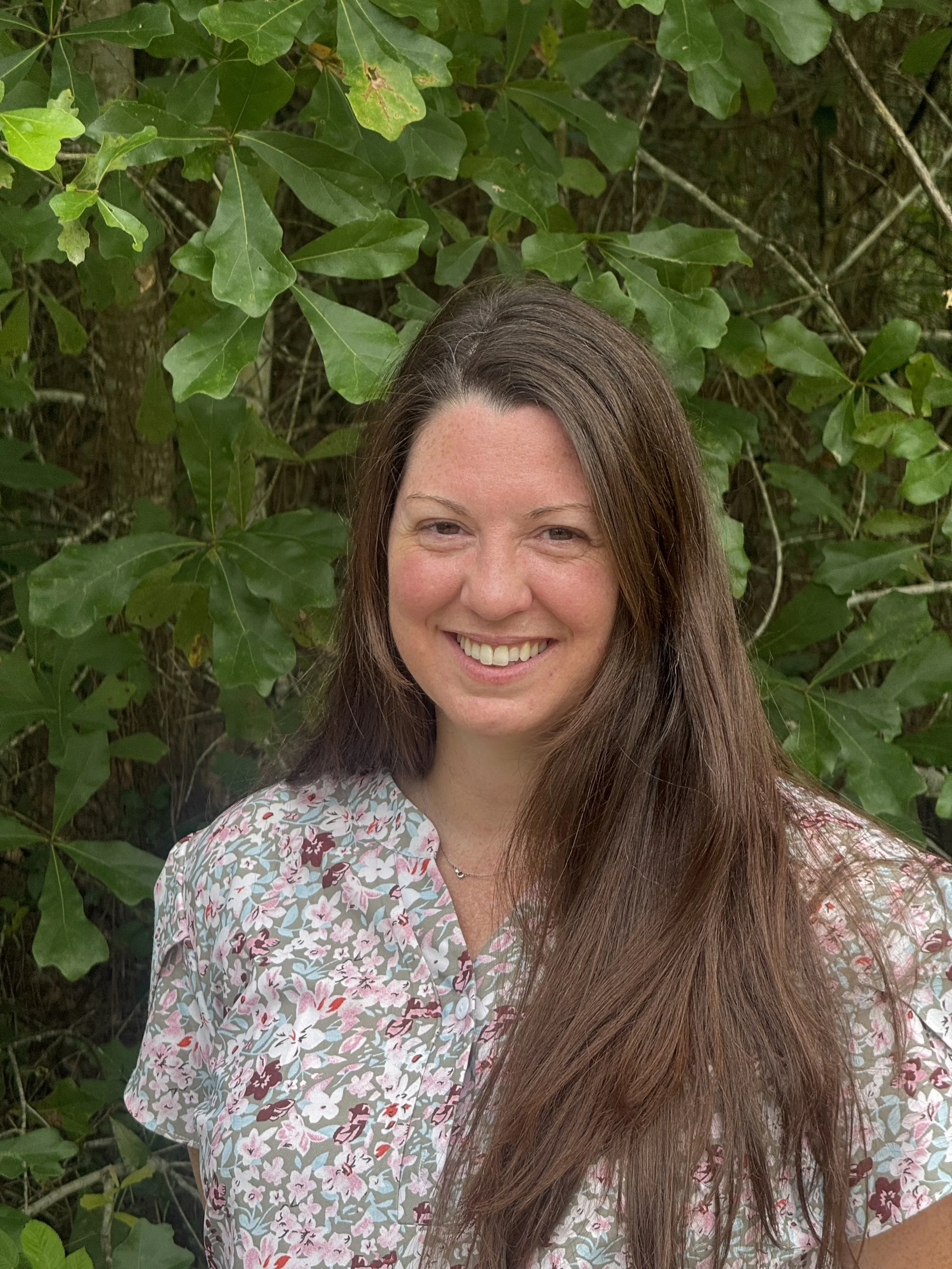 Tallecia, Veterinary Technician. A smiling woman with brown hair and freckles standing in front of green leafy bushes wearing a floral patterned shirt.