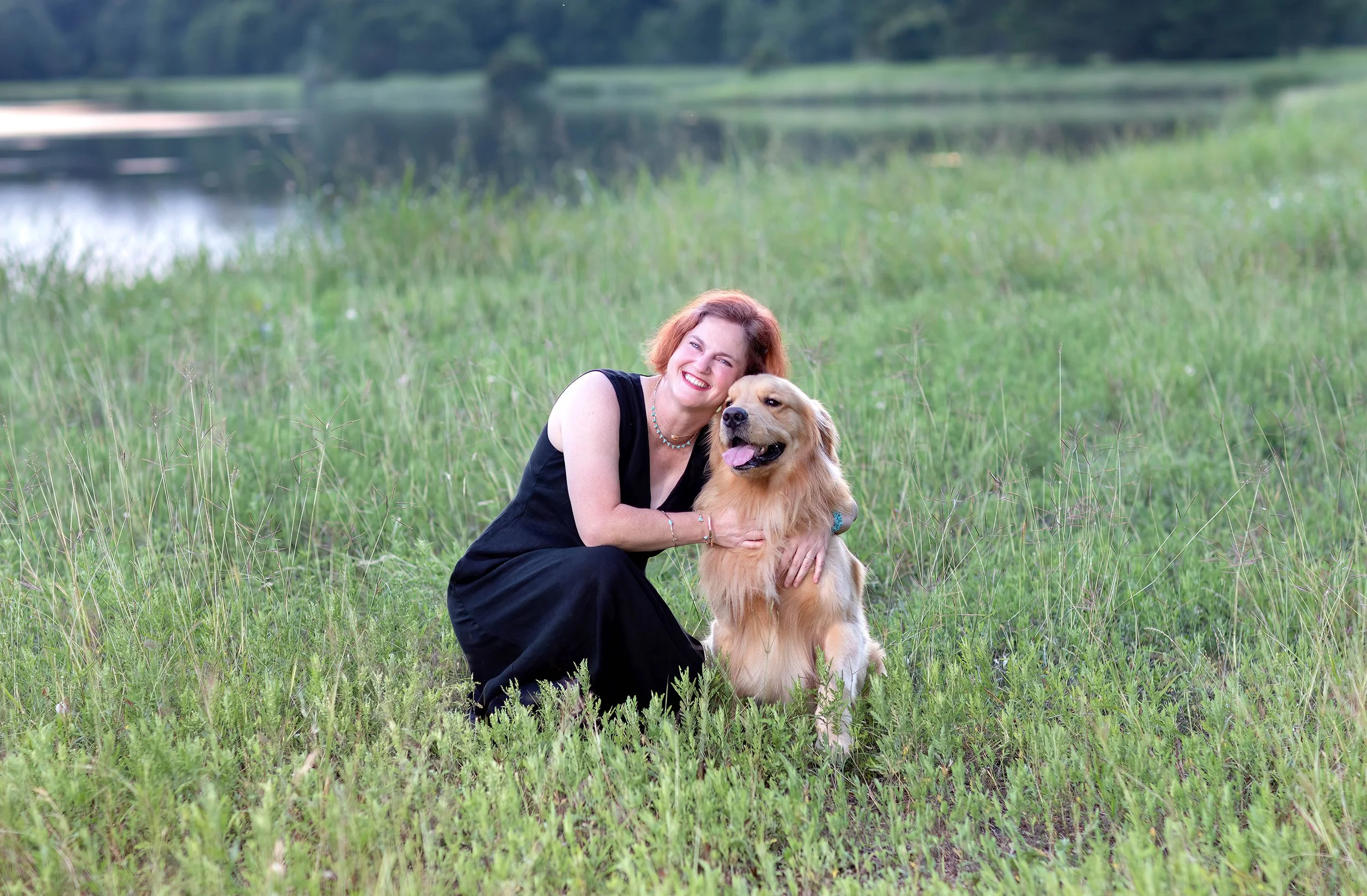 Dr. Bouche, A woman with short red hair in a black dress kneeling outdoors in a grassy field, smiling, hugging a large golden retriever dog with its tongue out near a lake with trees in the background in Cypress, Texas