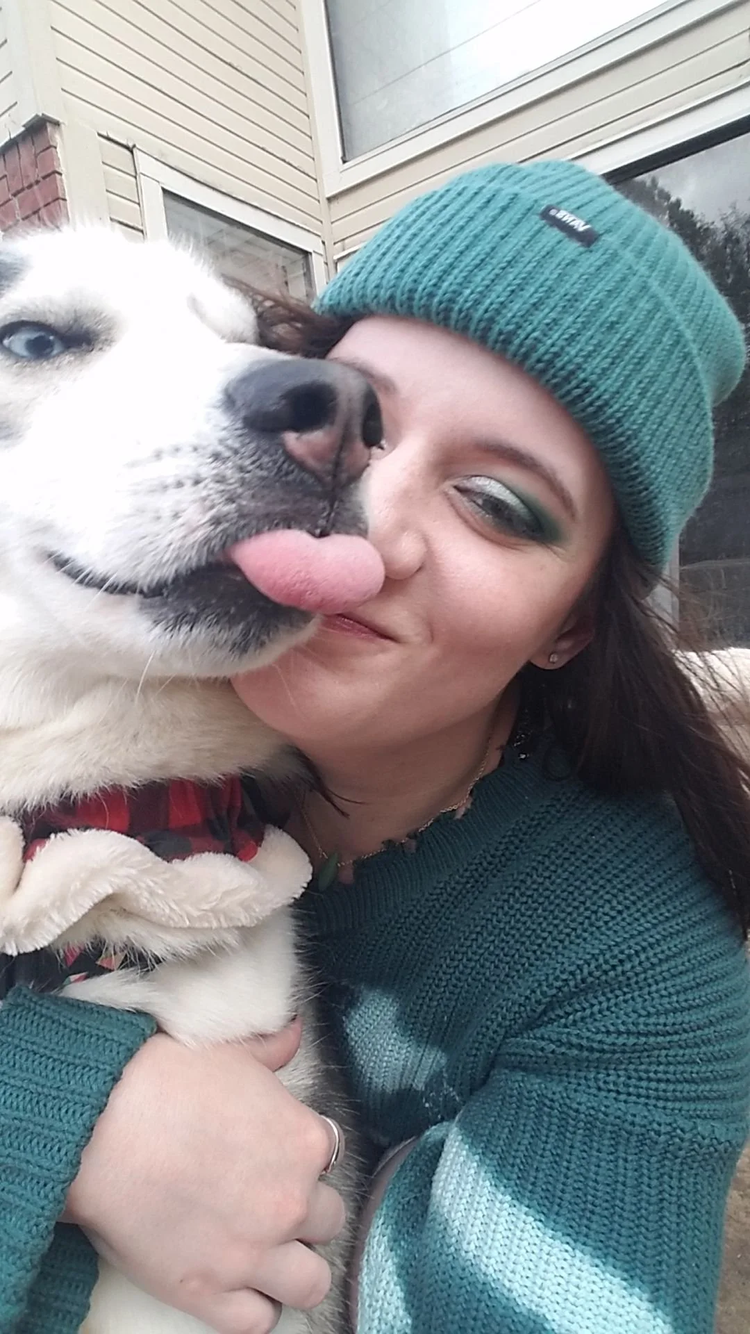 Katie, Client Care Coordinator. A woman with dark hair, wearing a teal knit beanie and green makeup, is smiling as she holds a husky dog close. The husky has light fur, blue eyes, and its tongue slightly sticking out.