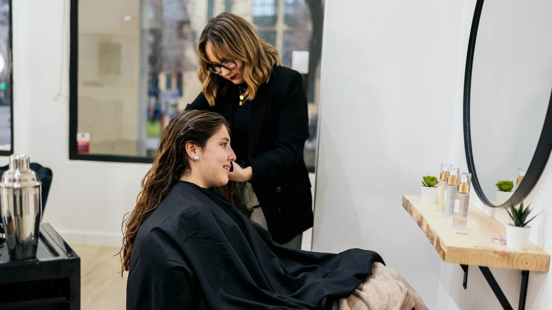 A woman with long brown hair and earrings is sitting in a salon chair, getting her hair styled. A hairstylist with glasses and shoulder-length hair is working on her hair. The salon has a window and a shelf with hair products and small potted plants.