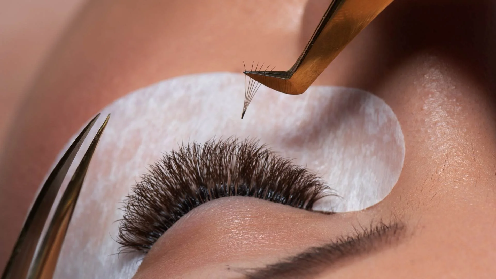 Close-up of a person receiving a lash extension procedure with tweezers, a surgical eye pad, and an eyelash at the center of the image.