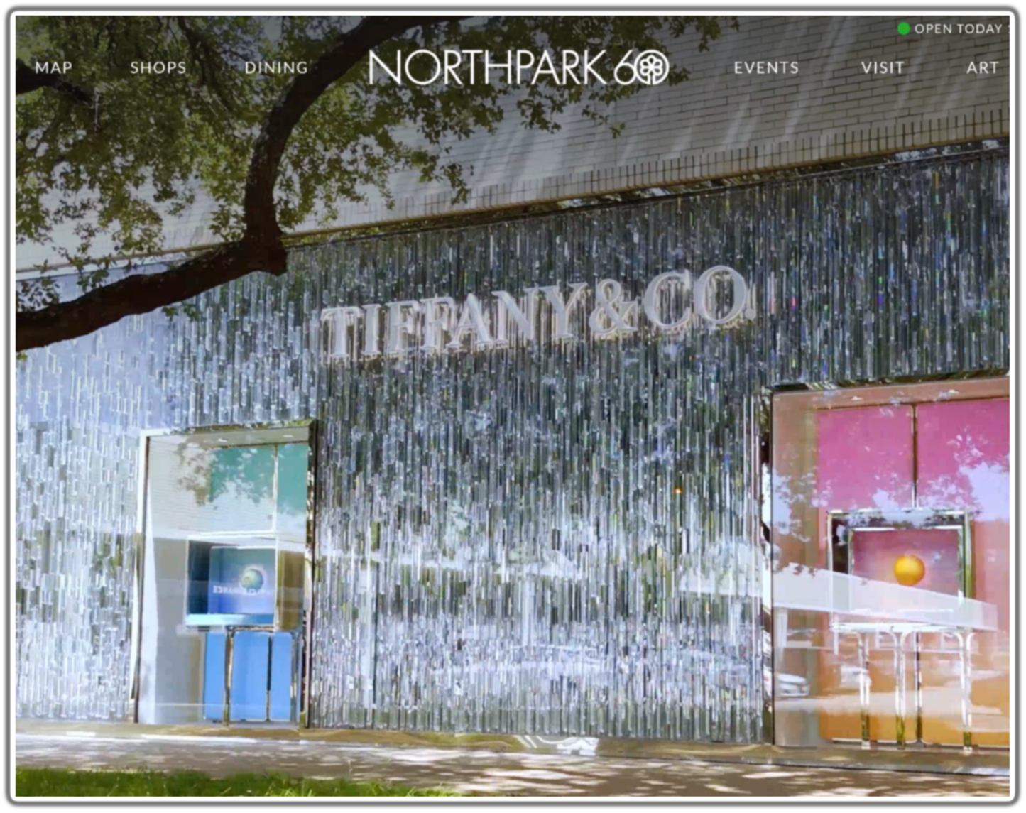 Facade of a Tiffany & Co. store with a shiny silver exterior and the store's logo, with trees reflected on the surface and a pink display window on the right.
