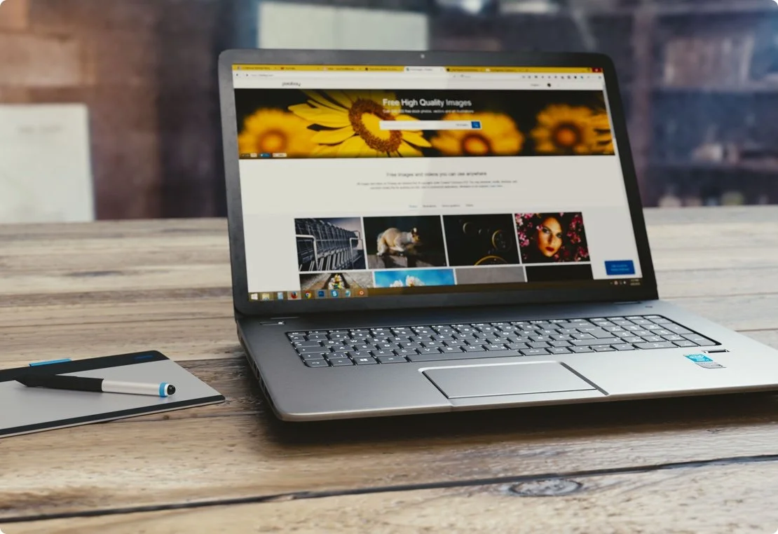 Laptop on a wooden table displaying a photo website, next to a black pen and a white notepad.